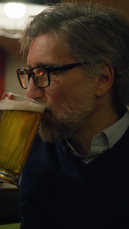 Vertical-shot-of-Caucasian-mature-man-drink-craft-beer,-talks-to-friend-and-bartender-sitting-at-the-bar-counter-in-stylish-pub-in-the-evening.-Male-friends-spend-weekend-away-from-home.-Lifestyle-and-leisure-concept.